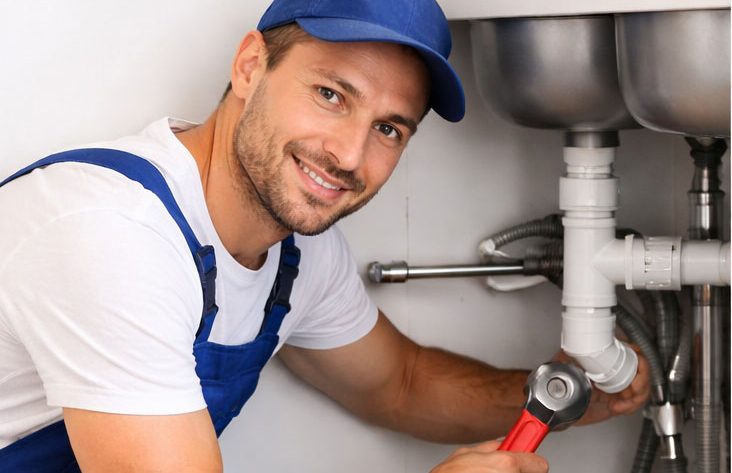 Lächelnder Klempner in blauer Arbeitskleidung arbeitet an einem Rohr.