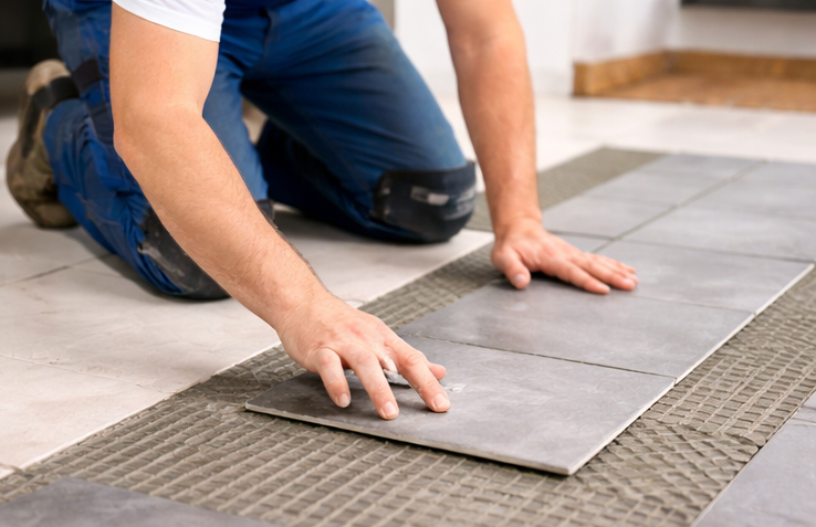 Handwerker verlegt graue Fliesen auf einem Bodenbelag.