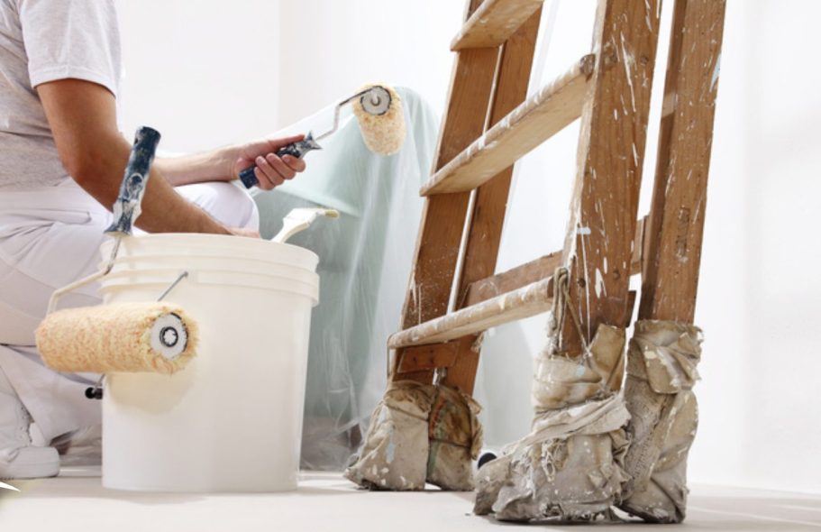 Ein Maler verwendet Farbroller und steht neben einer Leiter und einem Farbeimer.
